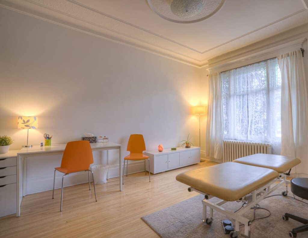 Bureau d'ostéopathie de Caroline Stoll Ostéopathe sur Montréal Le plateau, avec la table électrique, des chaises oranges, grande fenêtre, lumineuse